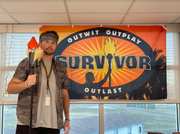 Garrett Betty (staff) dresses up and poses in front of a Survivor tapestry in his classroom. His classroom contains several items of Survivor memorabilia to show his interests.