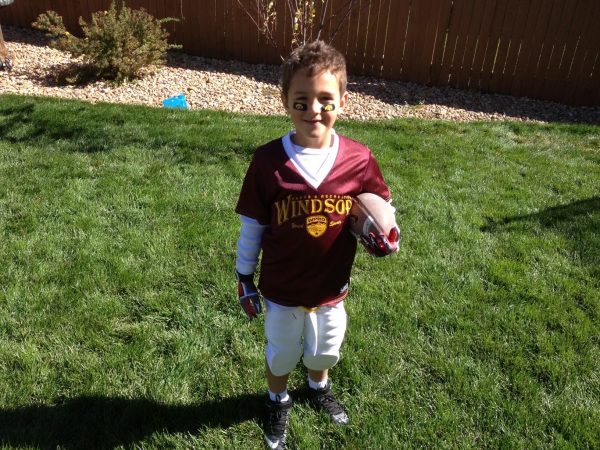 Little boy stands in a football uniform and smiles while holding a football